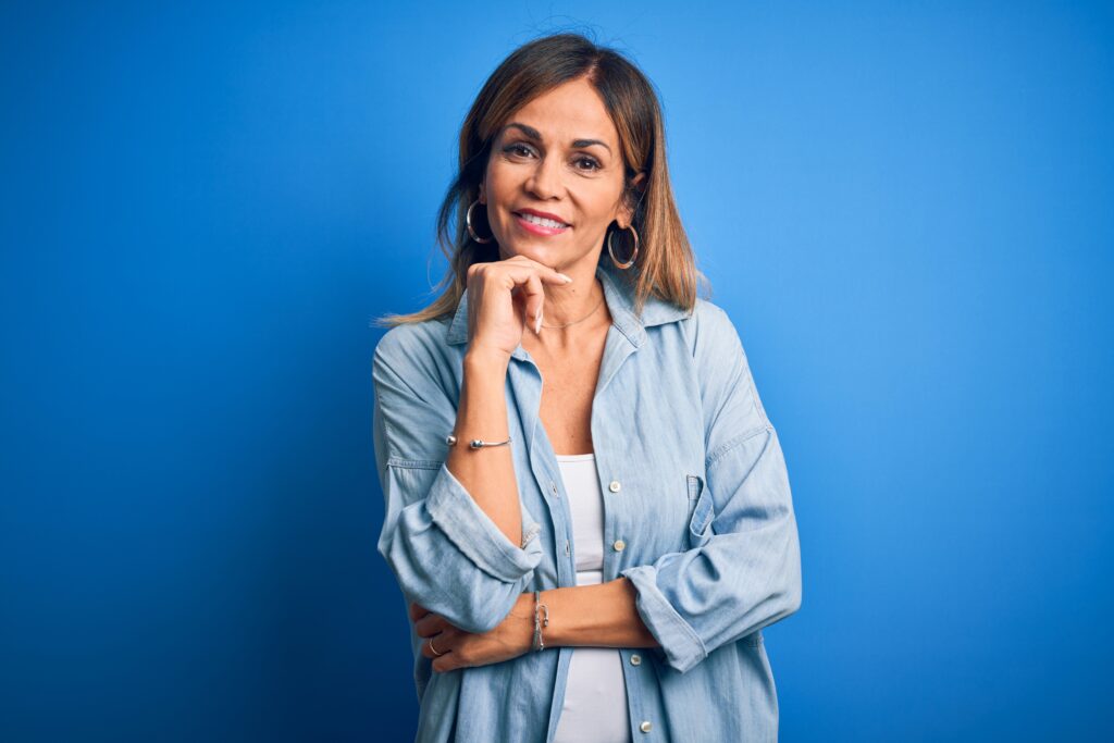 Middle aged woman smiling with perfect teeth, wearing casual shirt standing over isolated blue background looking confident at the camera smiling with crossed arms and hand raised on chin.